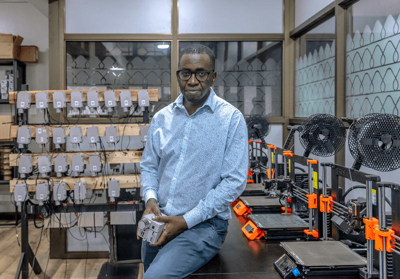 a black man sitting on a table surrounded by 3d printers and cables